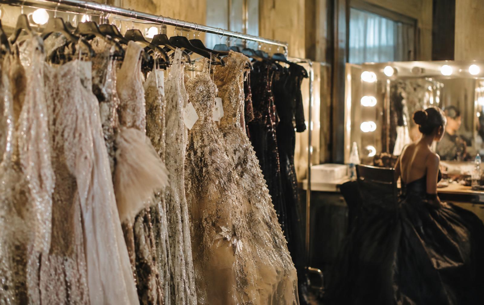 Rack of embellished couture gowns backstage beside a lit vanity mirror