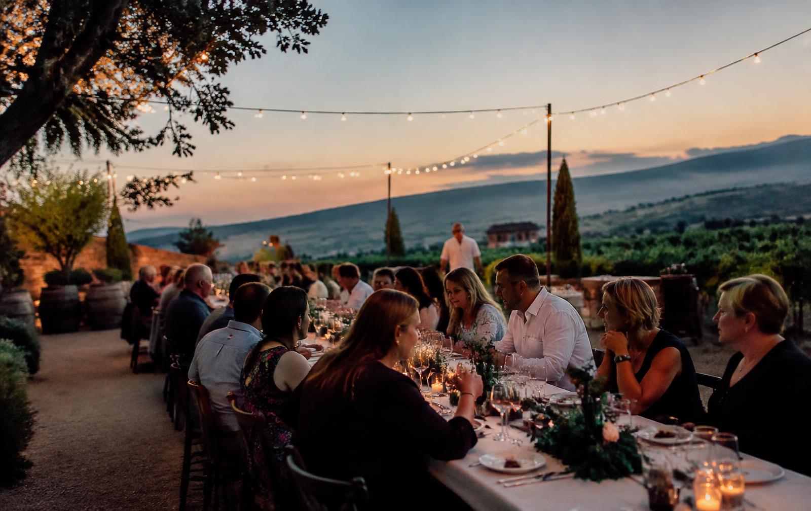 Travellers sharing a long-table dinner in a vineyard courtyard at dusk